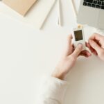 Person checking blood sugar with a glucometer on a white desk near a laptop.