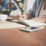 A woman writes in a notebook at a café table with a coffee and smartphone nearby.